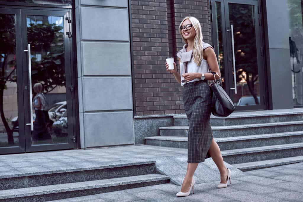 Female officer worker walking on street of downtown with morning coffee. Confident businesswoman in stylish formal wear looks at shop windows and holding coffee to go